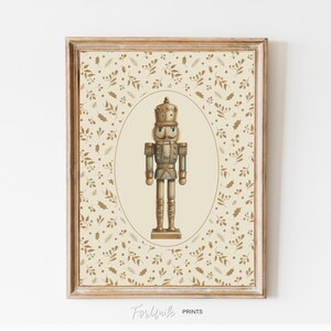 Könnte beinhalten: Ein gerahmter Druck mit einer Illustration im Vintage-Stil eines Nussknackersoldaten. Der Nussknacker ist in einem detaillierten, antiken Stil dargestellt, mit einer goldenen Krone und Uniform. Der Hintergrund ist cremefarben mit einem goldenen Blättermuster. Der Druck ist in einem goldfarbenen Holzrahmen gerahmt.