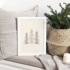 Puede incluir: Una impresi&oacute;n enmarcada en blanco con una ilustraci&oacute;n minimalista de tres &aacute;rboles estilizados en tonos verdes y marrones sobre un fondo crema. La impresi&oacute;n se muestra en un banco blanco con una manta de lana gris y una cesta de mimbre llena de ramas verdes.