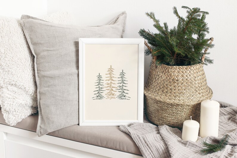 Puede incluir: Una impresi&oacute;n enmarcada en blanco con una ilustraci&oacute;n minimalista de tres &aacute;rboles estilizados en tonos verdes y marrones sobre un fondo crema. La impresi&oacute;n se muestra en un banco blanco con una manta de lana gris y una cesta de mimbre llena de ramas verdes.