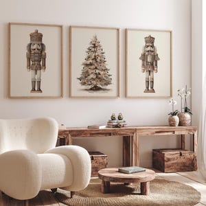 May include: Three framed art prints featuring a Christmas theme. The prints depict a nutcracker soldier, a Christmas tree, and another nutcracker soldier. The prints are in neutral tones and are displayed above a wooden console.