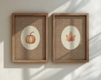 Peut inclure: Deux impressions encadrées sur le thème de l'automne. L'une représente une citrouille à l'aquarelle, l'autre une feuille d'érable, toutes deux sur un ovale beige sur un fond marron à motifs. Les cadres sont en bois brun clair.