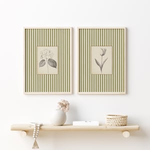 May include: Two framed botanical prints featuring a hydrangea and a tulip. The prints are in a cream-colored frame with a green and white striped background. The prints are displayed on a wooden shelf with a vase, beads, and a basket.