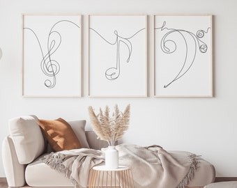 Könnte beinhalten: Drei minimalistische Schwarz-Weiß-Linienkunstdrucke von Musiknoten, einem Violinschlüssel und einem Bassschlüssel. Die Drucke sind in einem hellen Holzrahmen gerahmt und an einer weißen Wand über einem beigen Sofa mit einer Wolldecke aufgehängt.