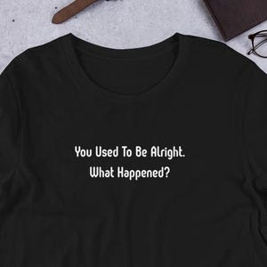 Puede incluir: Una camiseta negra de manga corta con el texto impreso en blanco "You Used To Be Alright. What Happened?" centrado en el pecho. La camiseta tiene un cuello redondo y está confeccionada con una tela negra sólida con letras blancas.