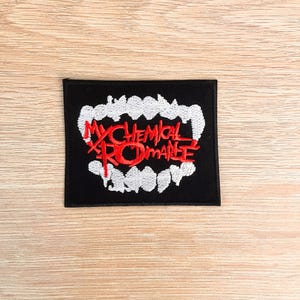 May include: A black embroidered patch with the words "MY CHEMICAL ROMANCE" in red, and a white tooth design. The rectangular patch is displayed on a light wooden surface.