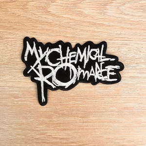 Peut inclure: Écusson brodé noir et blanc avec l'inscription "My Chemical Romance" dans une police stylisée. Le patch est bordé de noir et posé sur un fond en bois clair. Idéal pour personnaliser des vêtements ou des accessoires.
