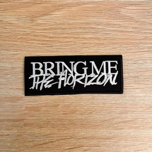 May include: A rectangular black embroidered patch featuring the white text BRING ME THE HORIZON. The top line displays the words BRING ME in a clean, bold font, while the bottom line features THE HORIZON in a stylised, jagged, hand-drawn script.