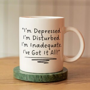 May include: White ceramic mug with the text "I'm Depressed. I'm Disturbed. I'm Inadequate. I've Got It All!" in black lettering. The mug is on a round, green marble coaster.