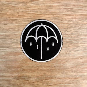 May include: A circular embroidered patch featuring a black background with a white stitched outline of an umbrella and four raindrops. The design is enclosed within a thin white circular border, creating a high-contrast graphic emblem.