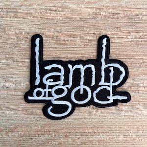 Puede incluir: Un parche bordado negro con las palabras "Lamb of God" en blanco, con una tipografía estilizada. El parche tiene forma rectangular con dos extensiones verticales en la parte superior. Ideal para ropa o accesorios.
