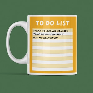 May include: A white ceramic mug with a yellow to-do list design. The list includes "Speak to ground control", "Take my protein pills", and "Put my helmet on".