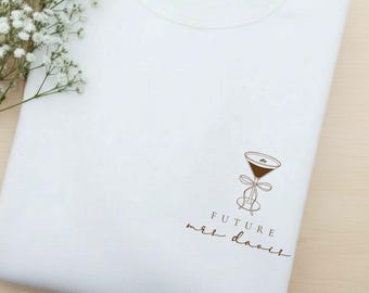May include: A white sweatshirt with a brown cocktail glass graphic and the text "FUTURE mrs. davis". The sweatshirt is folded on a light wooden surface with white flowers.