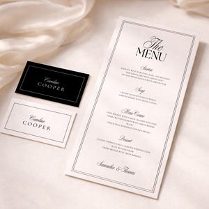 May include: Elegant wedding stationery set featuring a menu and two name cards. The menu, with a double border, lists courses like filet mignon and vanilla almond cake. The name cards display "Caroline Cooper" in a classic font. The background is a soft, white fabric.
