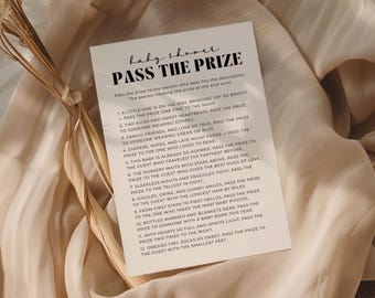 May include: A rectangular card with the text "baby shower PASS THE PRIZE" in black. The card includes a list of prompts for a baby shower game. The card is on a beige fabric background with dried plants.