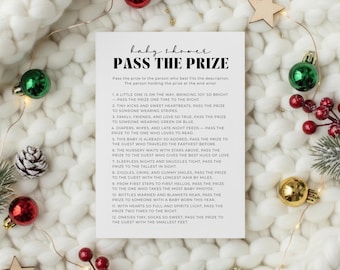 May include: A white card with the text "baby shower PASS THE PRIZE" sits on a white knitted blanket, surrounded by Christmas ornaments and lights. The card lists descriptions for a baby shower game.