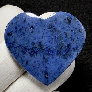 May include: A heart-shaped stone with a deep blue hue, speckled with darker blue and black inclusions. The polished surface reflects light, highlighting the natural patterns within the stone. The heart is held against a white cloth.