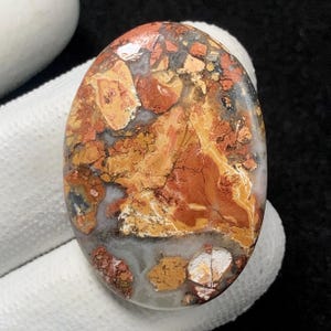 May include: Oval-shaped gemstone with a mosaic of colors. The stone features a blend of rusty red, golden yellow, and gray hues, creating a unique pattern. The polished surface highlights the natural variations and textures of the stone.