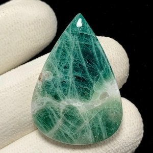 May include: A teardrop-shaped gemstone featuring a deep forest green color with intricate white veining throughout the surface. The stone has a smooth, polished finish with a reflective sheen and a pointed top edge, showcasing natural mineral patterns.