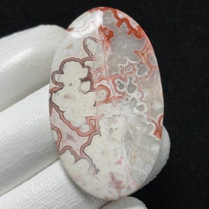 Puede incluir: Gema ovalada con un patrón único. La piedra presenta una base blanquecina con intrincados patrones arremolinados en tonos rojos y marrones. La superficie pulida resalta las variaciones y texturas naturales de la piedra.