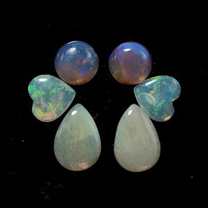 May include: A collection of polished opal gemstones in various shapes. The assortment includes round, heart, and teardrop shapes, displaying iridescent colors of blue, green, and pink against a black background. These gemstones are ideal for jewelry making.