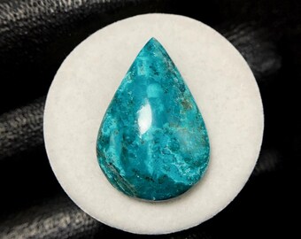 May include: A teardrop-shaped gemstone with a vibrant teal color and a polished surface. The stone is set on a white, circular base, with a dark background. The gemstone's unique color and shape make it a striking piece.