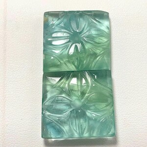 May include: Two rectangular, translucent gemstones with carved floral designs. The stones are a blend of blue and green hues, with intricate details. The carvings are visible on the surface of the stones.