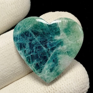 Puede incluir: Una piedra preciosa en forma de corazón con una superficie pulida que presenta un degradado de azul verdoso intenso a verde menta claro. La piedra muestra una compleja red interna de inclusiones cristalinas blancas y fracturas.