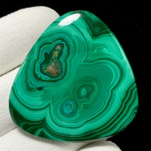 Puede incluir: Una piedra preciosa de malaquita pulida, de forma triangular. La piedra presenta bandas concéntricas de color verde oscuro y claro, creando un patrón arremolinado. Una zona central muestra una mezcla de tonos marrón y turquesa. El fondo es oscuro.