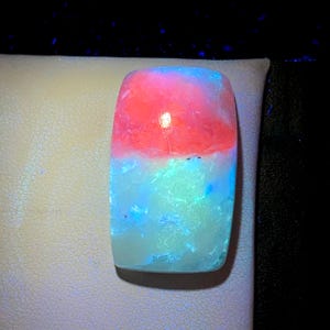May include: A rectangular cabochon gemstone displaying intense fluorescence under ultraviolet light. The top half glows with a vibrant neon pink hue, while the bottom half exhibits a bright, luminous cyan blue colour with a textured, crystalline surface.