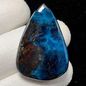 May include: A teardrop-shaped gemstone with a polished surface. The stone features a striking combination of deep blue and brown hues, with intricate patterns and textures. The gemstone is held against a white fabric.