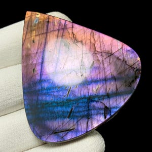 May include: A polished labradorite gemstone with a vibrant display of colors. The stone features a mix of purple, blue, and pink hues, with a pearlescent sheen. The surface has natural patterns and lines, creating a unique and eye-catching appearance.