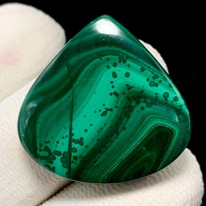 May include: A polished malachite gemstone, shaped like a teardrop. The stone is a deep green with swirling patterns and darker green spots. The surface is smooth and reflective, highlighting the natural beauty of the mineral. A natural gemstone.