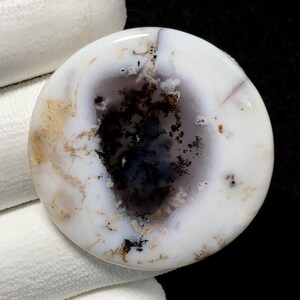 Puede incluir: Una gema redonda y pulida con una base blanca y patrones intrincados. La piedra presenta un área oscura central con inclusiones negras y azules, rodeada de vetas más claras, de color marrón claro. El efecto general es un diseño natural y orgánico.