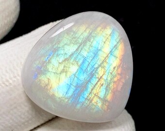 May include: A polished, oval-shaped moonstone gemstone. The stone is primarily white with iridescent flashes of blue, yellow, and orange light across its surface. The gemstone is set against a dark background.