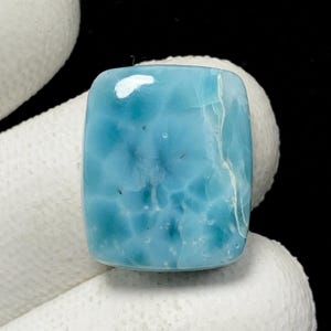 May include: A rectangular, polished gemstone with a light blue hue and a cloudy, textured appearance. The stone has a rounded top and a white inclusion on the upper left side. The gemstone is held by a white surface.