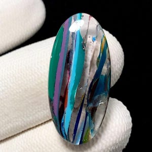 Può includere: Cabochon in vetro ovale con base trasparente e strisce verticali. Le strisce sono nelle tonalità di verde, viola, blu, verde acqua e argento. Il vetro ha una finitura lucida. Il cabochon è tenuto da una mano guantata.