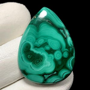 Puede incluir: Una piedra preciosa de malaquita pulida en forma de lágrima. La piedra presenta un color verde vibrante con patrones arremolinados de verde claro y oscuro, creando un diseño natural único. La piedra preciosa está sobre un fondo blanco.