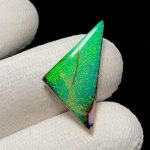 May include: A triangular-shaped gemstone featuring a vibrant, iridescent green surface with a fine, sparkling texture and dark, thin linear inclusions running across the face, displaying subtle hints of purple along one of the edges.