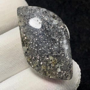 Könnte beinhalten: Ein polierter, tropfenförmiger Edelstein mit einem gesprenkelten, glitzernden Aussehen. Der Stein weist eine Mischung aus grauen, schwarzen und silbernen Flecken auf, mit einem prominenten weißen Einschluss in der Nähe der Mitte. Der Edelstein wird vor einem weißen Stoff gehalten.