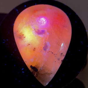 Puede incluir: Una gema en forma de lágrima con un gradiente de color vibrante. La piedra pasa de un naranja cálido a un amarillo brillante, con toques de púrpura e inclusiones negras. La gema se encuentra sobre un fondo oscuro, resaltando sus cualidades luminosas.