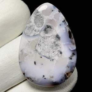Puede incluir: Una gema pulida en forma de lágrima. La piedra presenta una mezcla de patrones blancos, grises y negros, creando un efecto veteado único. La paleta de colores es suave y neutra, con un acabado brillante.