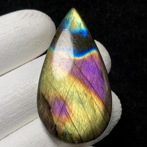 May include: A teardrop-shaped labradorite gemstone with a polished surface. The stone displays a vibrant play of color, including iridescent blues, purples, and yellows. The gemstone is held by a gloved hand against a dark background.