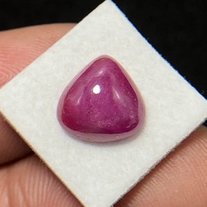 Puede incluir: Una piedra preciosa de rubí pulida, tallada en cabujón. La gema es de un color rojo-rosa profundo y saturado, con una forma triangular redondeada y lisa. Se encuentra sobre una superficie blanca cuadrada, con un fondo oscuro.