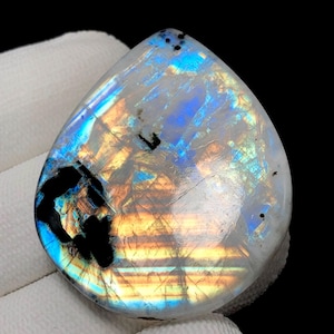 May include: A pear-shaped, polished gemstone with a white base and iridescent flashes of blue, orange, and yellow. The stone has dark, irregular inclusions and a smooth, reflective surface. The gemstone is set against a white background.