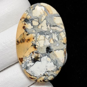 May include: An oval-shaped gemstone with a unique pattern. The stone features a blend of colors, including beige, cream, gray, and black, creating a mosaic-like appearance. The surface is polished, highlighting the natural variations and textures.