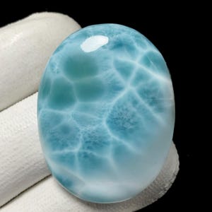 May include: A polished, oval gemstone in shades of light blue, featuring a distinctive web-like pattern of white veins. The cabochon has a glossy finish and is set against a dark backdrop. Ideal for jewellery, the stone is a cabochon cut.