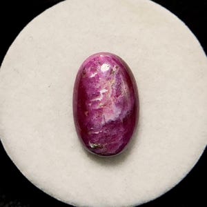 Könnte beinhalten: Ovaler rosafarbener Rubin-Edelstein mit weißen Streifen und einer glatten, polierten Oberfläche.
