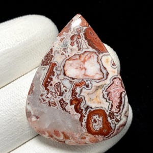 May include: A teardrop-shaped gemstone with a unique pattern of red, brown, white, and pink swirls and veins. The polished stone is held against a white surface, showcasing its natural beauty and intricate design.