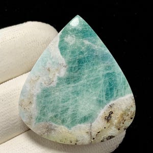 May include: A teardrop-shaped gemstone with a polished surface. The stone features a blend of light teal and white hues, with areas of beige and dark speckling. The overall appearance suggests a natural, organic material, possibly a polished stone or mineral.