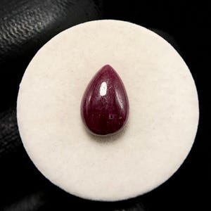 May include: A deep red, pear-shaped gemstone with a smooth, polished cabochon surface. The stone displays a rich, saturated crimson hue and a single prominent light reflection on its rounded top, centered on a circular white display base.
